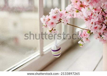 Happy Easter!Beautiful branches of cherry blossoms.A bouquet of pink flowers.A decorative purple chicken egg on a ribbon.Home decor on a white window in spring for a holiday .Copy Space Royalty-Free Stock Photo #2133897265