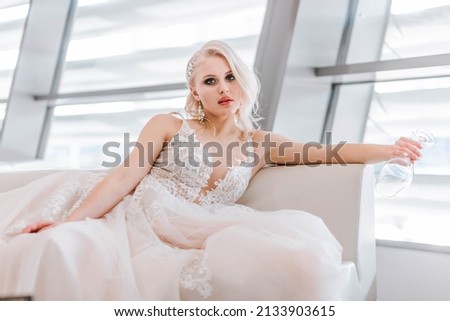 An attractive pensive blonde woman with a creative hairstyle for short hair  is sitting on a white sofa in the lobby of the hotel. Art photography. White long wedding dress of the bride. 