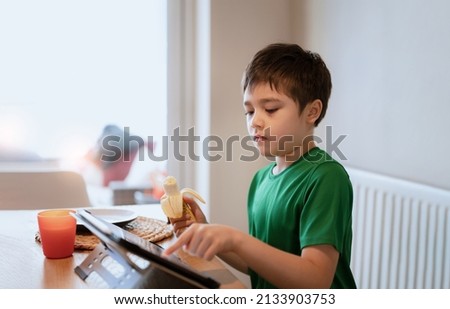 Child boy eating banana and watching cartoon on tablet, Kid having  breakfast and playing game online on internet with friends before go to school in the morning