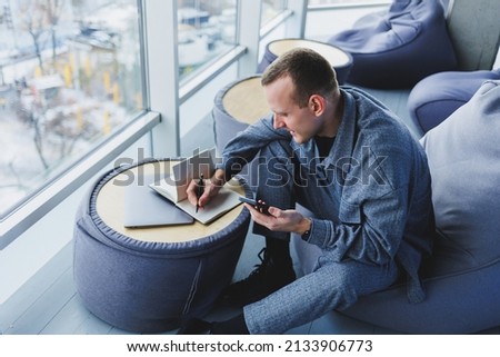 A handsome European young businessman in a coffee shop sits with a mobile phone and makes notes in a notebook. Wearing an elegant casual suit and sitting with a laptop in a public cafe