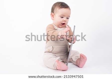 Adorable baby boy wearing beige overalls sitting on white background holding a tablet pad and watching streaming videos or cartoons. 