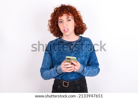 Photo of astonished crazy young redhead girl wearing blue sweater over white background hold smartphone dislike feedback concept