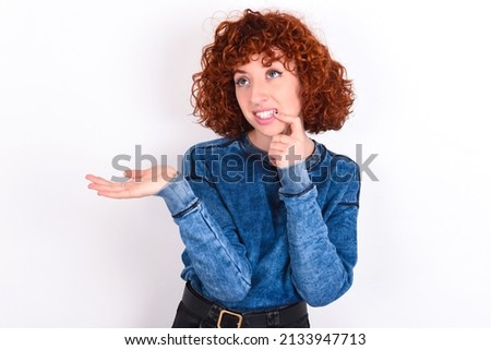 Positive young redhead girl wearing blue sweater over white background advert promo touch finger teeth