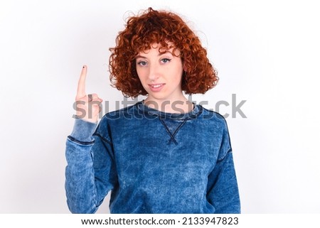 young redhead girl wearing blue sweater over white background smiling and looking friendly, showing number one or first with hand forward, counting down