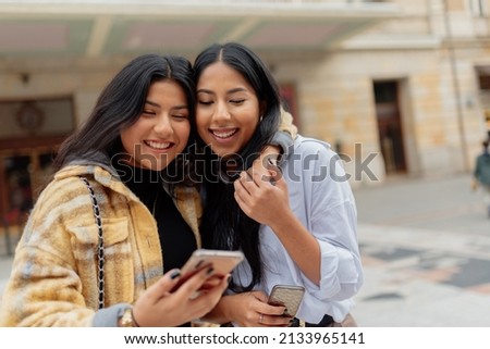 Two Latin women looking at each other and holding their mobile phones in the street. Hispanic sister having a good time together sightseeing. Royalty-Free Stock Photo #2133965141