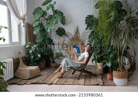 Stress relief after work from home. Relaxed young lady spend time in cozy indoor garden with monstera plant sitting with closed eyes and blowing fan. Love for plants, wellness and wellbeing concept Royalty-Free Stock Photo #2133980361