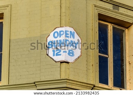 An old metal sign with the message “open Sunday 12 -8” attached to a painted brick wall