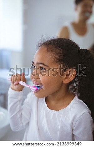 Multiracial girl brushing teeth while looking away with father in background. unaltered, family, togetherness and hygiene concept. Royalty-Free Stock Photo #2133999349