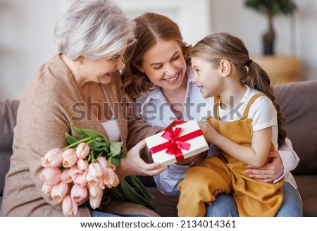 Happy International Women's Day. Smiling multi generational family daughter and granddaughter giving flowers  and gift to grandmother  cheerfully celebrate Mother's Day or grandparents day at home