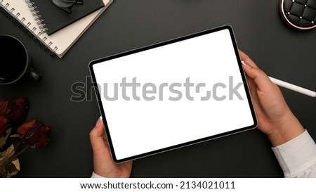 Top view, A businesswoman hands holding tablet touchpad white screen mockup over modern black office desk with office accessories.