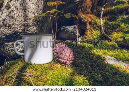 Enamel white mug on the mossy ground with cedar cone forest mockup. Trekking merchandise and camping geer marketing photo. Stock wildwood photo with white metal cup. Rustic scene, product template.