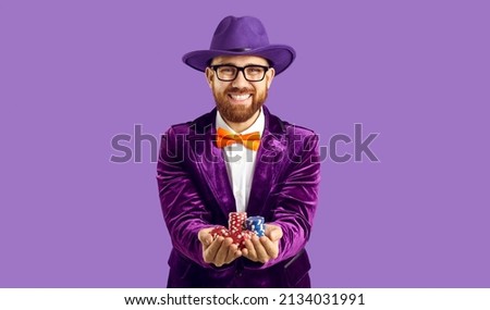Portrait of smiling male entertainer in suit and hat isolated on purple studio background hold casino chips. Happy man player or poker professional isolated on purple studio background. Royalty-Free Stock Photo #2134031991