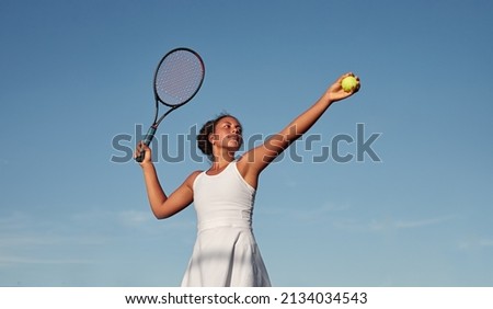 From below African American female athlete in white dress preparing to hit ball with racket against blue sky while playing tennis in daytime Royalty-Free Stock Photo #2134034543