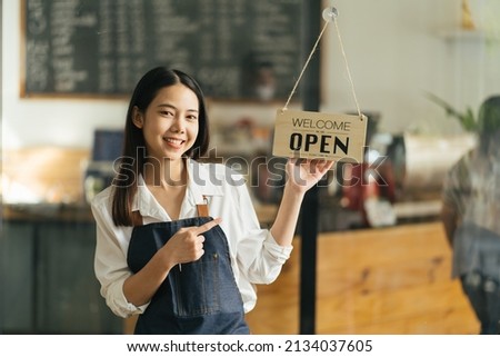 Asian Happy business woman is a waitress in an apron, the owner of the cafe stands at the door with a sign Open waiting for customers. Small business concept, cafes, and restaurants