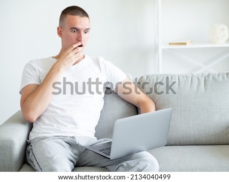 Shocked news. Emotional man. Unexpected information. Surprised expressive casual guy sitting sofa looking laptop on knees in light room interior copy space. Royalty-Free Stock Photo #2134040499