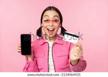 Asian girl shows mobile phone screen and credit card, reacts surprised at camera, gasping impressed, standing over pink background, online shopping concept