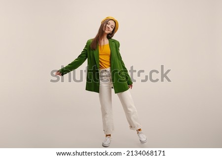 In full growth, cheerful young caucasian brunette girl poses against backdrop in studio. Teenager wears green jacket, white pants and yellow beret. Lifestyle, beauty concept Royalty-Free Stock Photo #2134068171