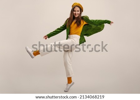 Full length young caucasian girl moves vigorously waving legs on white background in studio. Brunette wears yellow beret, blouse, green jacket and pants. Good mood, fashion trends Royalty-Free Stock Photo #2134068219