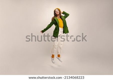 Happy young caucasian woman in beret, jacket and trousers is jumping in studio on white background. Brunette has fun at photoshoot. People sincere emotions lifestyle concept.