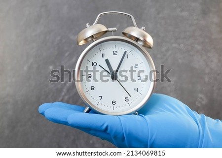 alarm clock held by a hand in a medical glove on a gray background.