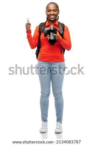people, profession and photography concept - happy smiling woman photographer with digital camera and backpack showing thumbs up over white background