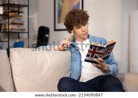 A handsome college student reads an English dictionary while sitting on the couch in the living room of his family's home. A teenage boy in a denim shirt repeats more new words in his mind. Royalty-Free Stock Photo #2134090847