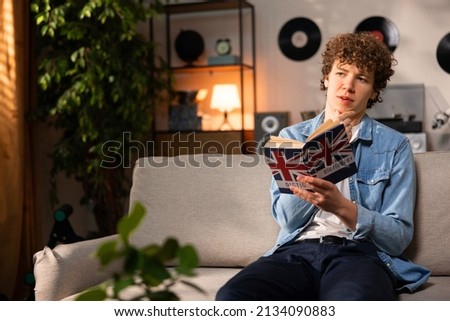 A handsome college student reads an English dictionary while sitting on the couch in the living room of his family's home. A teenage boy in a denim shirt repeats more new words in his mind. Royalty-Free Stock Photo #2134090883