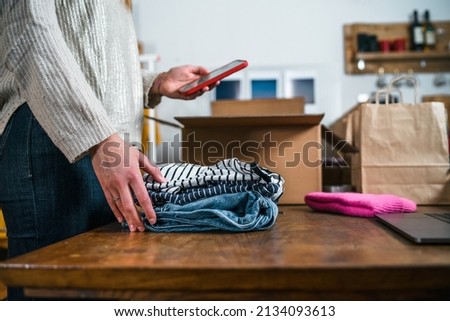 Business woman prepares a parcel for shipping in a cardboard box with clothes from her online store - Millennial sells second-hand used dresses in her home using the smartphone application - Start up Royalty-Free Stock Photo #2134093613
