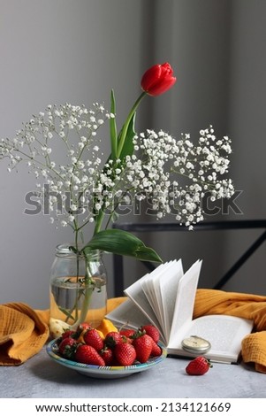 Red tulip in glass jar, strawberry on a plate, an open book on a table. Summer still life with fruit. 