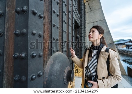 asian lady holding camera is admiring and touching the giant heavy gate in Japan. chinese female tourist standing by entrance is lost in thought while feeling the ancient metal gate. Royalty-Free Stock Photo #2134164299
