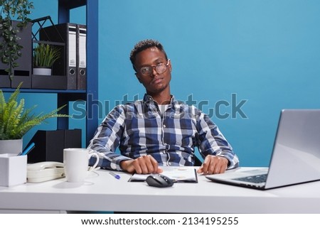 Bored and tired young adult business person sitting at office desk while looking at camera. Exhausted and fatigued agency employee with boredom face expression sitting in workspace interior.