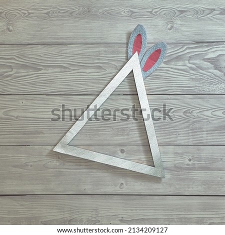 Triangular silver frame and bunny ears on a light gray wooden background. Text space. Top view. Minimal style. Easter.