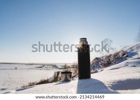 A black thermos of tea stands on the snow behind the sun. Outdoor recreation during a winter hike, a mug with mustard tea in nature. High quality photo