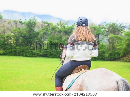 Back view of a caucasian woman wearing an andean sweater and riding a horse in the meadow. Horizontal. With copy space. Royalty-Free Stock Photo #2134219589