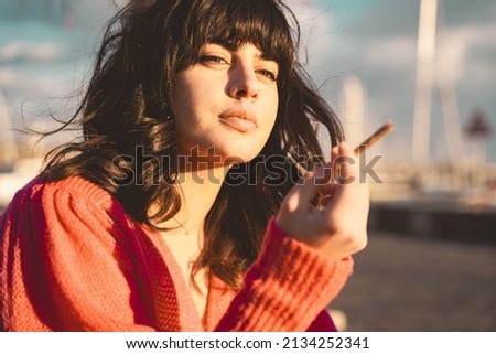 Beautiful brunette smoking a rolled cigarette outdoors sitting at the harbor bench against the sunset sun
