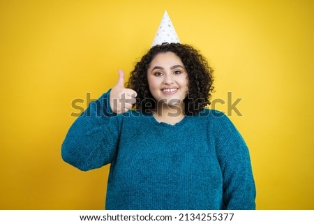 Young beautiful woman wearing a birthday hat over isolated yellow background success sign doing positive gesture with hand, thumb up smiling and happy. cheerful expression