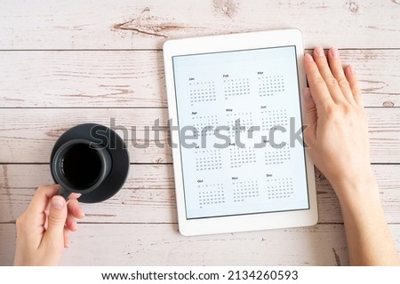 tablet computer with an open app of calendar for unspecified unknown date year without date and cup of coffe or tea in a womans hands on a wooden boards background. top view, flat lay