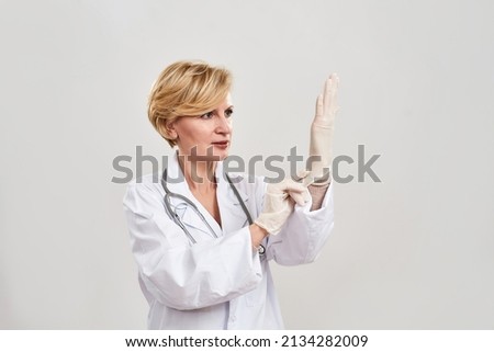 Adult female doctor putting latex glove on her hand. Caucasian woman with stethoscope wearing white coat. Concept of medical and health care. Isolated on white background. Studio shoot. Copy space Royalty-Free Stock Photo #2134282009