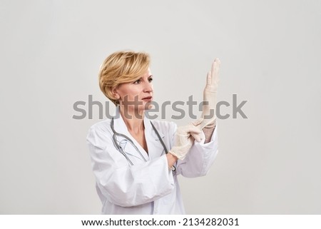 Mature female doctor putting latex glove on her hand. European woman with stethoscope wearing white coat. Concept of medical and health care. Isolated on white background. Studio shoot. Copy space Royalty-Free Stock Photo #2134282031