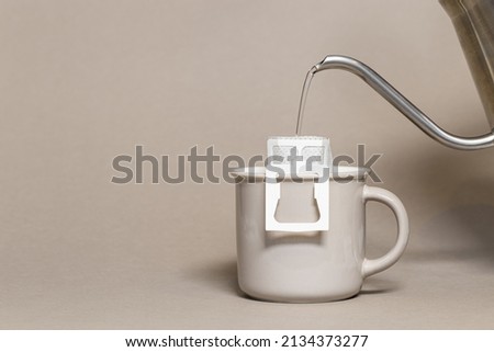 Drip coffee bag with ground coffee in big cup, hot water is poured into paper filter drip coffee for brewing on beige color background with copy space, quick way to brew ground coffee Royalty-Free Stock Photo #2134373277