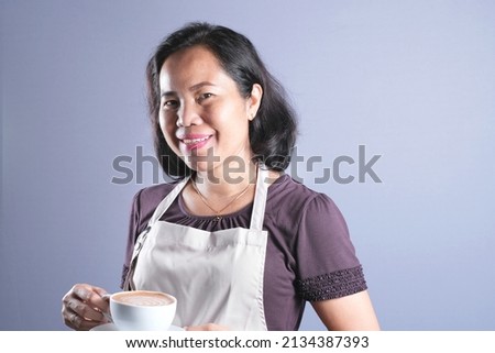 Asian housewife woman barista looks happy wearing apron holding a cup of coffee isolated over grey background. Copy space.