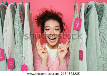 Young fun costumer woman wear sweater stand near clothes rack with tag sale in store showroom waiting for special moment, keeping fingers crossed, making wish isolated on plain pink background studio