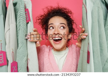 Close up young fun smiling minded happy caucasian female costumer woman 20s wearing sweater stand near clothes rack with tag sale in store showroom isolated on plain pink background studio portrait