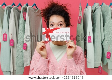 Young woman in sweater stand near clothes rack with tag sale in store showroom cover face hold gift certificate coupon voucher card for store do winner gesture isolated on plain pink background studio