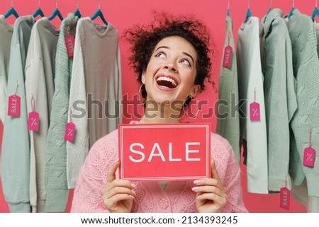 Young minded female costumer woman wear sweater stand near clothes rack with tag sale in store showroom hold card sign with sale title text look aside isolated on plain pink background studio portrait