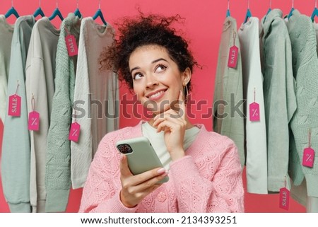 Young minded wistful dreamful female costumer woman wear sweater stand near clothes rack with tag sale in store showroom hold use mobile cell phone isolated on plain pink background studio portrait