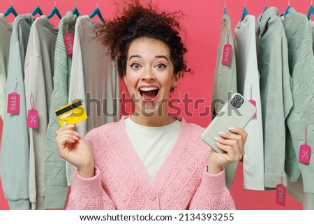 Young excited amazed woman in sweater stand near clothes rack with tag sale in store showroom using mobile cell phone hold credit bank card shopping online isolated on plain pink background studio.