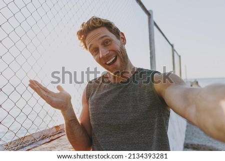 Young strong sporty athletic fit sportsman man in sports clothes warm up train do selfie shot mobile cell phone spread hand at sunrise sun over sea beach outdoor on pier seaside in summer day morning