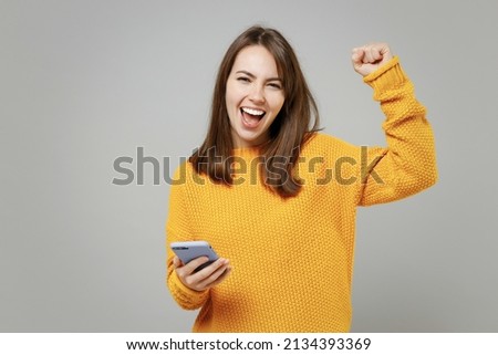 Young smiling cheerful woman 20s wearing casual knitted yellow sweater hold in hand using mobile cell phone chatting browsing typing sms messaging isolated on grey color background studio portrait. Royalty-Free Stock Photo #2134393369