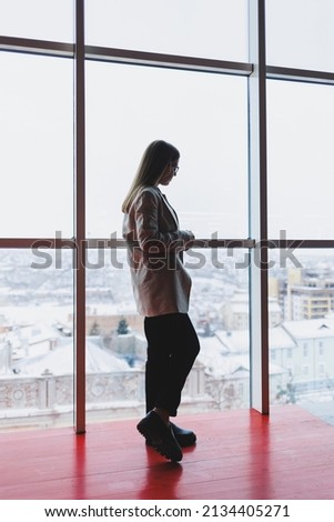 A successful business woman in trousers and a jacket stands and looks out the huge panoramic window of the business center skyscraper.